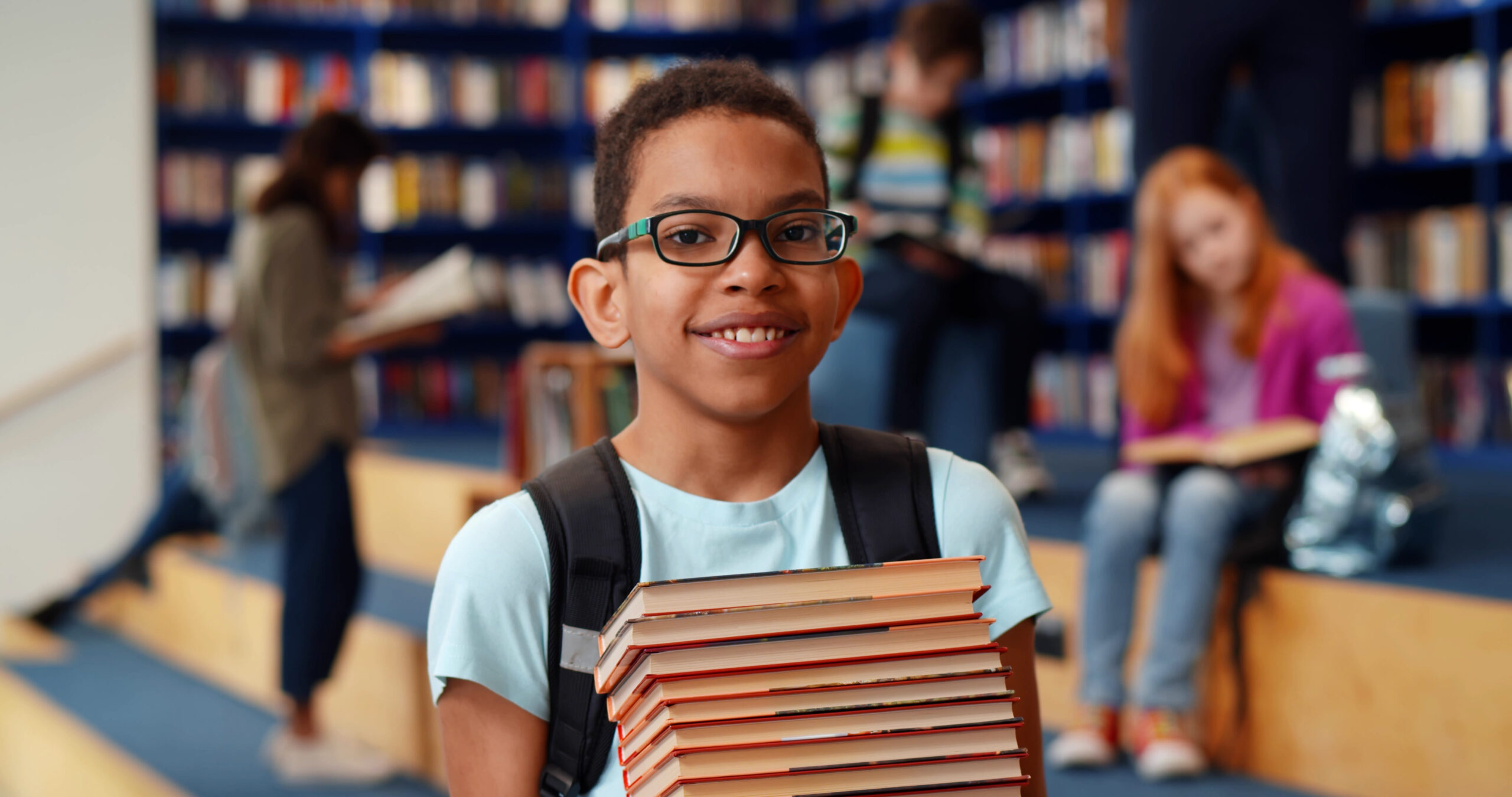 Middle eastern boy holding stack of books against multi colored bookshelf in library.